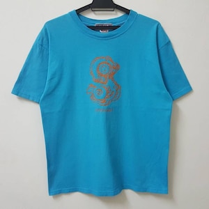 以下が含まれることがあります： ターコイズブルーのTシャツに、茶色のスタイリッシュなタコと「Seaquest」の文字が描かれています。