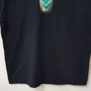 K&ouml;nnte beinhalten: Schwarzes Kurzarm-T-Shirt mit einem farbenfrohen Grafikdesign auf der Vorderseite. Die Grafik scheint ein stilisiertes Bild eines gr&uuml;n-goldenen Objekts zu sein. Das Shirt ist aus einem weichen, dunklen Stoff gefertigt.