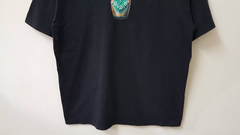 K&ouml;nnte beinhalten: Schwarzes Kurzarm-T-Shirt mit einem farbenfrohen Grafikdesign auf der Vorderseite. Die Grafik scheint ein stilisiertes Bild eines gr&uuml;n-goldenen Objekts zu sein. Das Shirt ist aus einem weichen, dunklen Stoff gefertigt.