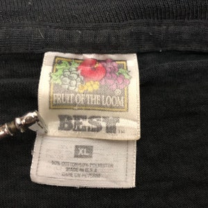 May include: A white tag with black text that reads "Fruit of the Loom" and "Best" with a small image of fruit. The tag is attached to a black t-shirt.