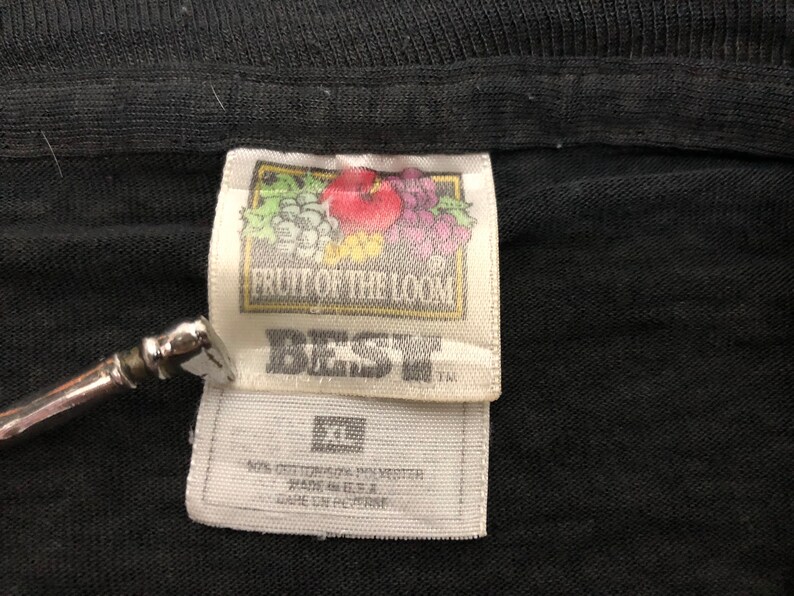 May include: A white tag with black text that reads "Fruit of the Loom" and "Best" with a small image of fruit. The tag is attached to a black t-shirt.