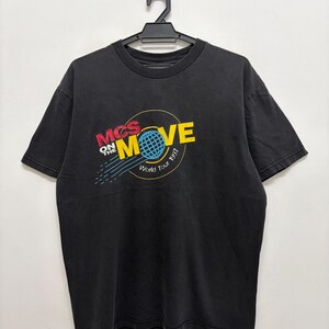 Op de afbeelding: Zwart t-shirt met een kleurrijke afbeelding. De afbeelding toont de woorden "MCS ON THE MOVE" in rood en geel, met een blauwe wereldbol in het midden. Onder de wereldbol staat de tekst "World Tour 1997".