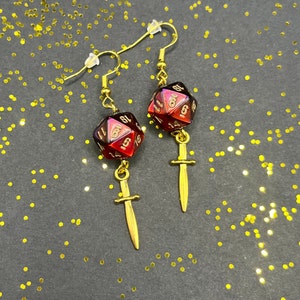 Peut inclure: Une paire de boucles d'oreilles en or avec des breloques de dés rouges irisés et des pendentifs d'épée dorés.