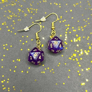 Peut inclure: Une paire de boucles d'oreilles pendantes dorées avec des breloques de dés violets irisés. Les dés ont des chiffres dorés sur chaque face.