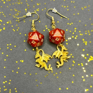 Peut inclure: Une paire de boucles d'oreilles en forme de dragon dorées avec des breloques de dés rouges, dorés et blancs. Les dés ont les chiffres de 1 à 12.