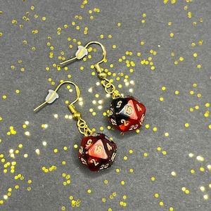 Peut inclure: Une paire de boucles d'oreilles en or avec des breloques de dés rouges et noirs. Les dés ont des chiffres dorés.
