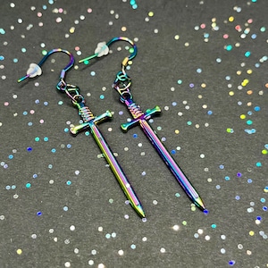 Peut inclure: Une paire de boucles d'oreilles en forme d'épée arc-en-ciel irisées. Les épées sont détaillées avec une garde croisée et une pointe pointue. Les boucles d'oreilles sont en métal et ont une fermeture à crochet.