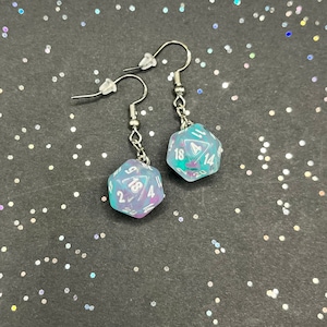 Peut inclure: Une paire de boucles d'oreilles pendantes argentées avec des breloques de dés bleu et violet irisés. Chaque dé a des chiffres blancs sur les faces.