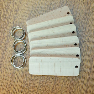 May include: Five unfinished wooden keychains with a hole at the top. The keychains are stacked on top of each other. The keychains are made of light wood with a natural grain.