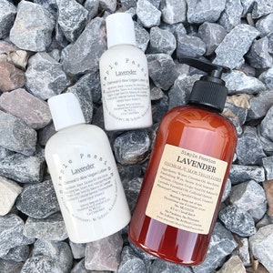 May include: Three bottles of Simple Passion Lavender Oatmeal & Aloe Vegan Lotion. The bottles are on a bed of gray rocks. The labels are white with black text and list the ingredients. The largest bottle is brown glass with a pump dispenser.