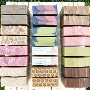 May include: A collection of 16 handmade bars of soap arranged in four rows of four. The soaps are in various colors and patterns, including brown, green, pink, white, and black. Some of the soaps have swirls, while others have a honeycomb pattern.