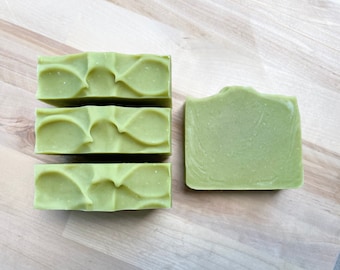 May include: Four bars of green soap, three stacked and one lying flat. The soap has a textured surface and is made with natural ingredients.