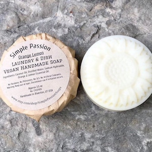 May include: A round, off-white vegan handmade soap with a sunflower design next to a wrapped bar. The wrapper reads "Simple Passion Orange Lemon Laundry & Dish Soap." The soap weighs approximately 3.5 oz.
