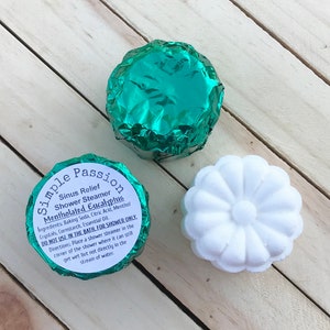 K&ouml;nnte beinhalten: Zwei in gr&uuml;nes Folienpapier verpackte Duschsteamer und ein wei&szlig;er Duschsteamer in Form einer Blume. Die gr&uuml;nen Steamer haben den Text "Simple Passion Sinus Relief Shower Steamer Mentholated Eucalyptus" auf dem Etikett. Das Etikett enth&auml;lt auch Gebrauchsanweisungen: "NICHT IM BAD VERWENDEN, NUR F&Uuml;R DIE DUSCHE. Anleitung: Legen Sie einen Duschsteamer in die Ecke der Dusche, wo er noch nass werden kann, aber nicht direkt im Wasserstrahl."