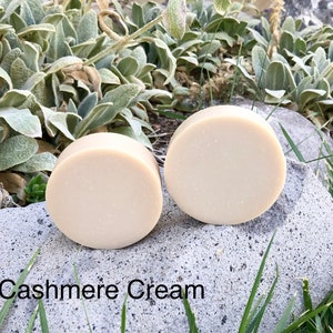 Könnte beinhalten: Zwei runde Stücke offweißer Seife, beschriftet mit "Cashmere Cream", liegen auf einem grauen Stein mit grünem Gras und Blättern im Hintergrund.