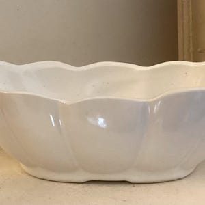 May include: A white, oval ceramic bowl with a scalloped edge. The bowl has a glossy finish and a simple design. It is likely used for serving food or as a decorative item. The bowl's dimensions are in centimetres.