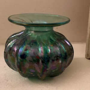 May include: A small, green glass vase with iridescent swirls and speckles. The vase has a wide, flat top and a rounded body.