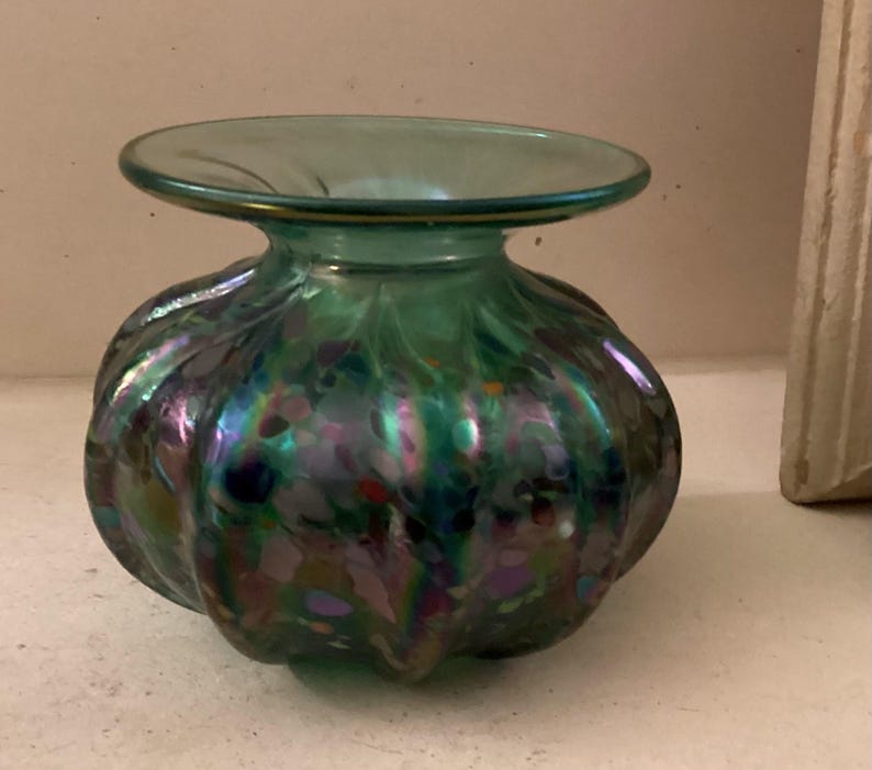 May include: A small, green glass vase with iridescent swirls and speckles. The vase has a wide, flat top and a rounded body.