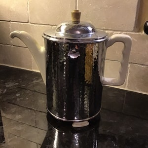 May include: A silver hammered metal coffee pot with a white porcelain spout and handle. The pot has a wooden knob on the lid.