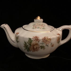 May include: White teapot with a gold trim and floral design. The teapot has a lid with the word "November" written on it.