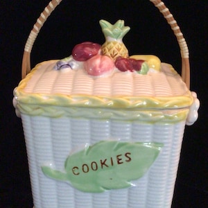 May include: White ceramic cookie jar shaped like a basket with a brown wicker handle. The lid is decorated with a pineapple and other fruit. The front of the jar has a green leaf with the word "COOKIES" written on it.
