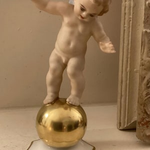 May include: A porcelain figurine of a cherub balancing on a gold sphere. The cherub has light skin, curly hair, and is nude. The sphere sits on a white, star-shaped base with gold trim. A decorative item.