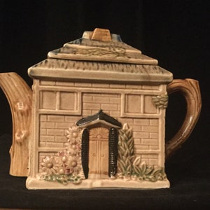 May include: A ceramic teapot shaped like a small house with a brown roof, brown brick walls, a brown door, and green plants. The teapot has a brown handle and spout.
