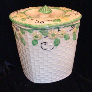 May include: White ceramic cookie jar with a green lid and floral design. The jar has a woven pattern and is decorated with yellow flowers and green leaves.
