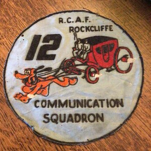 May include: A circular patch with the text "R.C.A.F. ROCKCLIFFE" and "COMMUNICATION SQUADRON". The patch features a cartoon dog pulling a red carriage with the number "12" in black.