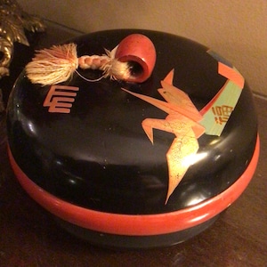 Peut inclure: Une boîte ronde laquée noire avec un bord et un couvercle rouges. Le couvercle présente un motif de grue et d'autres éléments décoratifs dans des tons orange et or. Une touffe et une poignée cylindrique rouge sont attachées au couvercle.