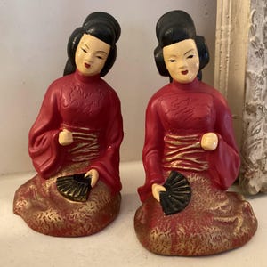 May include: Two ceramic figurines of women in red and gold robes holding black fans. The women are kneeling and have black hair.