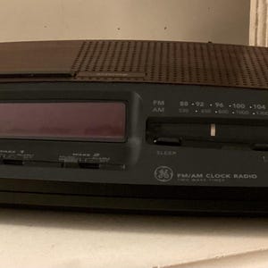 May include: A black GE FM/AM clock radio with a digital display. The radio has a brown speaker grill and a tuning dial. The clock radio has buttons for sleep, on, and off. The display shows the time in red.