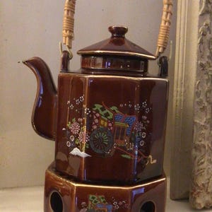 May include: A brown ceramic teapot with a wicker handle and a matching base. The teapot features a floral design with a Japanese-style cart and mountain scene.