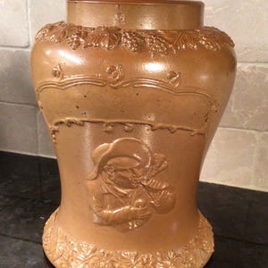 May include: A brown ceramic pitcher with a relief design of grapes and a man playing a musical instrument. The pitcher has a wide base and a narrow neck.