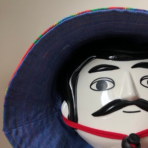 Può includere: Un cappello a secchiello blu e arcobaleno con fodera in denim blu e un cinturino regolabile rosso con fibbia nera.