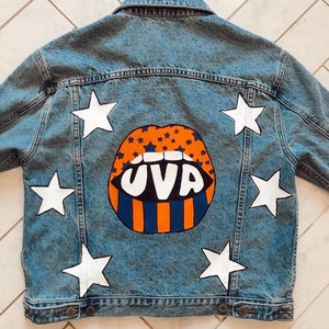 Puede incluir: Una chaqueta vaquera azul con estrellas blancas y unos labios pintados de naranja, azul y blanco con el texto "UVA" en la espalda.