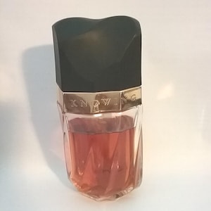 May include: A perfume bottle with a black, heart-shaped lid and a gold band that reads "KNOWING." The bottle contains a pink liquid. The glass bottle has a geometric design.