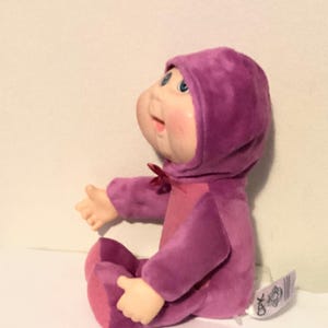 May include: A plush doll with a round face, blue eyes, and rosy cheeks. The doll is wearing a purple hooded outfit with a matching bow. The doll is sitting with its arms outstretched, and a tag is attached to its side.