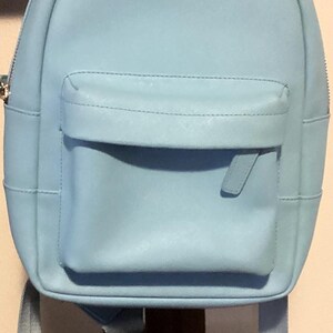 May include: A light blue backpack featuring a top handle, a main zippered compartment, and a front zippered pocket. The name ARIS is embroidered in silver thread on the upper front section of the bag, which also includes two adjustable shoulder straps.