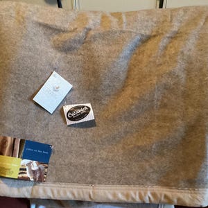 May include: A tan-colored blanket with a soft, felted texture and a finished hem. Attached are three tags: a white tag with a heart logo, an oval tag reading Creswick Woollen Mills, and a rectangular tag with an image of an alpaca and text.