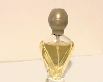 May include: A small, clear glass perfume bottle with a light yellow liquid inside. The bottle has a unique, stylized shape with a gray, rounded cap. The perfume bottle is sitting on a white surface.