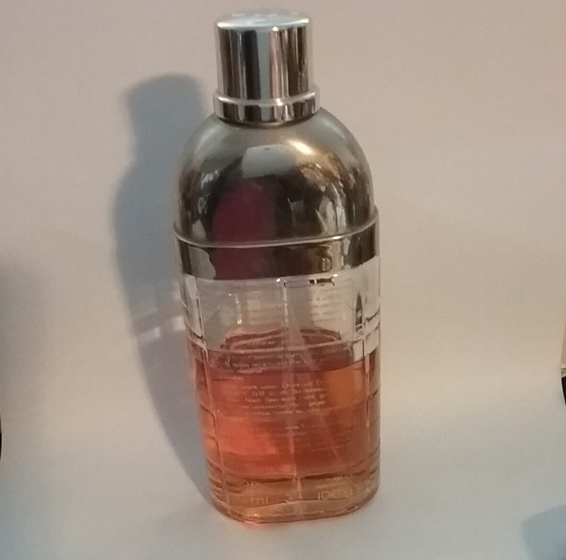 May include: A clear glass perfume bottle with a silver cap and a silver band around the middle. The bottle contains a pink liquid. The bottle is shaped like a cocktail shaker.