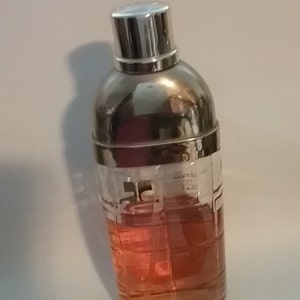 May include: A cocktail shaker with a silver top and a clear glass body filled with a reddish-orange liquid. The glass has a grid-like pattern. The top is shiny and reflective.