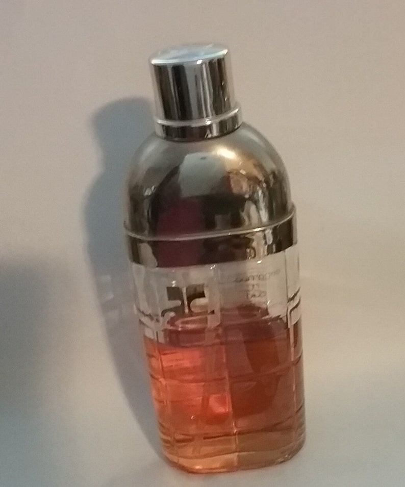 May include: A cocktail shaker with a silver top and a clear glass body filled with a reddish-orange liquid. The glass has a grid-like pattern. The top is shiny and reflective.