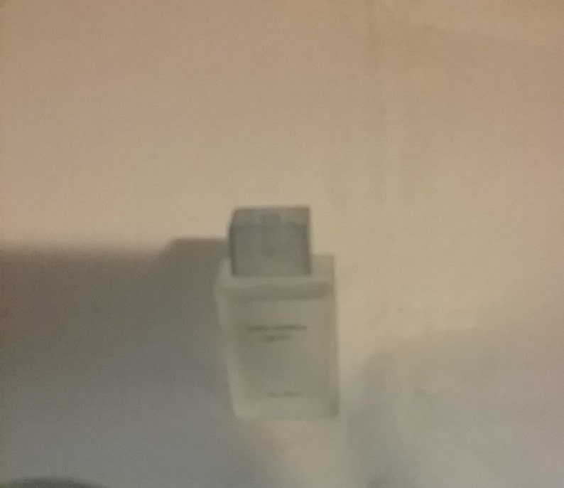 Puede incluir: Un frasco de perfume rectangular y transparente con tap&oacute;n plateado. El frasco tiene un dise&ntilde;o minimalista con texto en la parte delantera. El fondo es una pared lisa de color blanquecino.