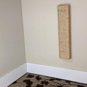 May include: A beige wall-mounted cat scratching post covered in natural sisal rope. The post is mounted in the corner of a room with white baseboards and a patterned carpet.