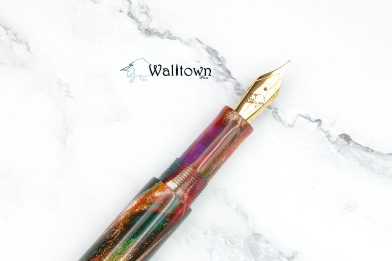 May include: A fountain pen with a gold nib and a multi-coloured body. The pen's body has a swirling pattern of red, green, purple, and gold. The pen is set against a white marble background. The brand name "Walltown" is visible.