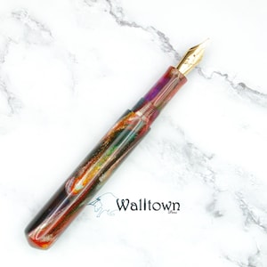 May include: A vibrant fountain pen with a gold nib. The pen's body has a marbled effect in shades of red, purple, green, and gold. It is displayed on a white marble surface.