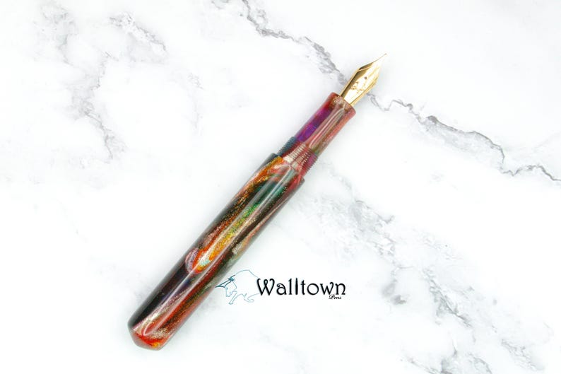 May include: A vibrant fountain pen with a gold nib. The pen's body has a marbled effect in shades of red, purple, green, and gold. It is displayed on a white marble surface.