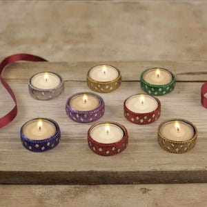 Könnte beinhalten: Eine Sammlung von brennenden Teelichtern in dekorativen Haltern. Die Halter sind in verschiedenen Farben gehalten, darunter Gold, Silber, Lila, Rot, Grün und Blau, mit einem Rautenmuster. Die Kerzen stehen auf einer Holzoberfläche.